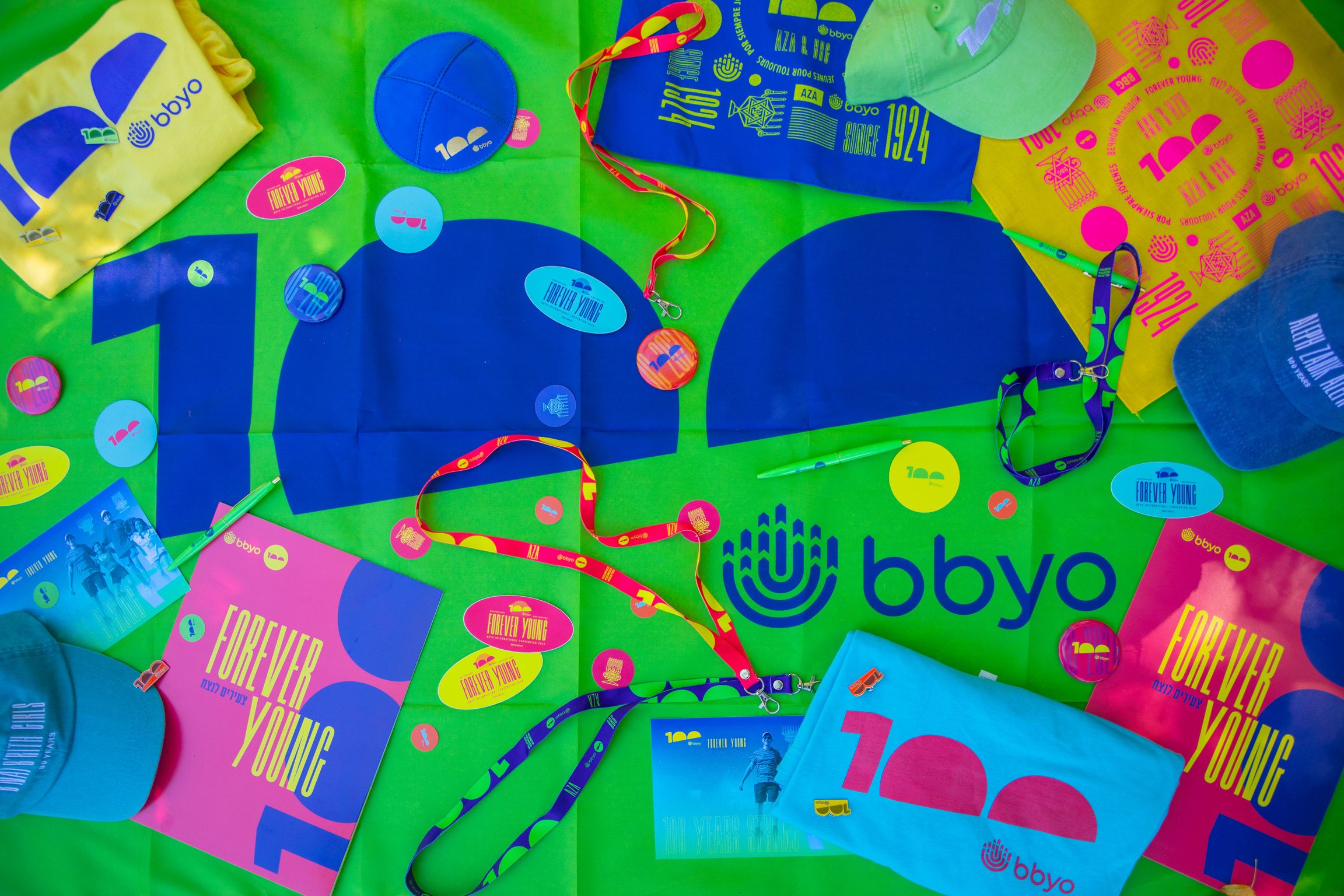 BBYO Centennial merch flatlay: lanyards, pins, hats, and programs